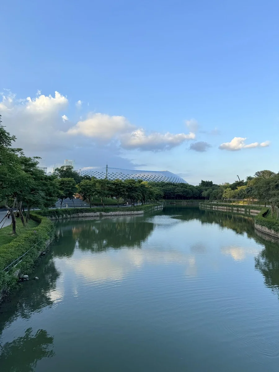 Calm canal in Rencai Park, Shenzhen, with the iconic diamond-patterned building and city skyline reflected in the water under