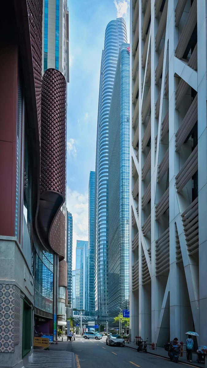 Towering glass skyscrapers frame a narrow urban street in Shenzhen's Futian CBD, with distinctive architectural details on ad