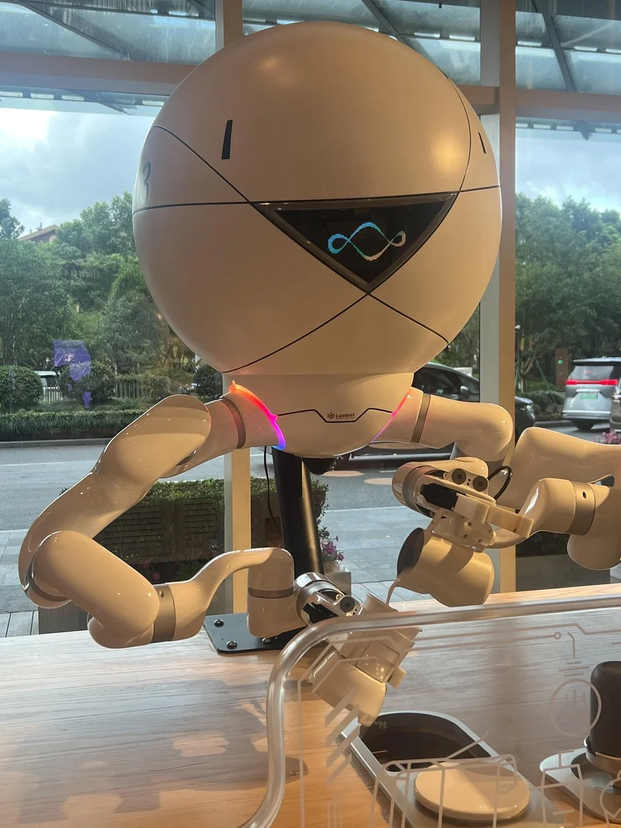 A white spherical robot with glowing arms and a triangular display is making coffee at a café counter, viewed through a windo