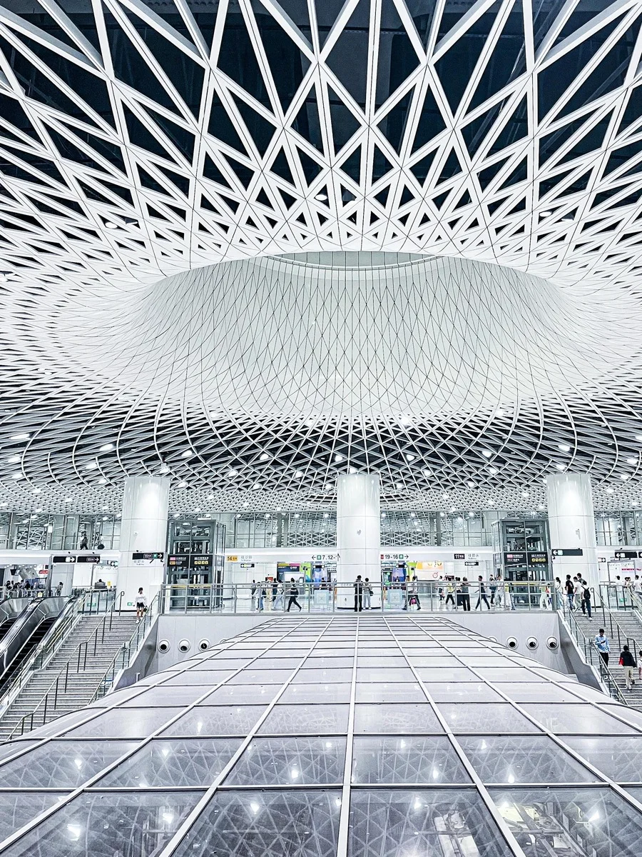Interior of Gangxia Bei Metro Station in Shenzhen, featuring a striking geometric white ceiling and glass walkway with commut