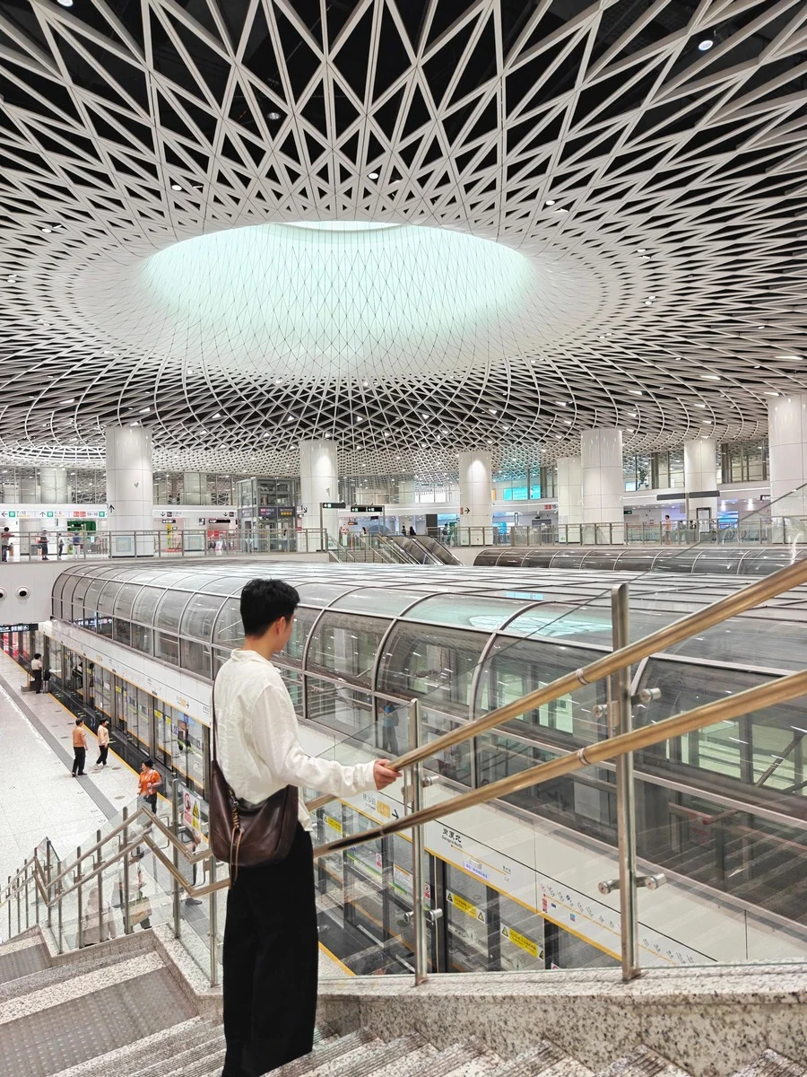 A person stands on stairs overlooking the futuristic, geometric ceiling of GuaXia Bei Metro Station in Shenzhen, with a train