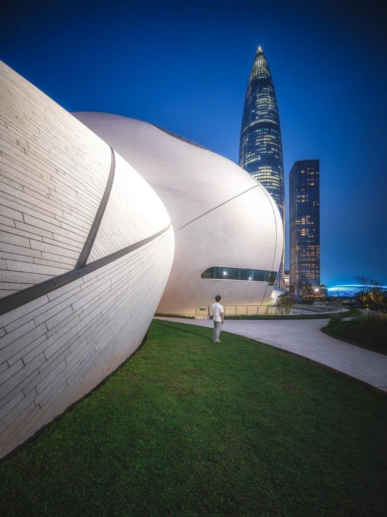 A person stands on a grassy slope facing the curvilinear stone facade of Shenzhen Bay Cultural Plaza at dusk, with the iconic