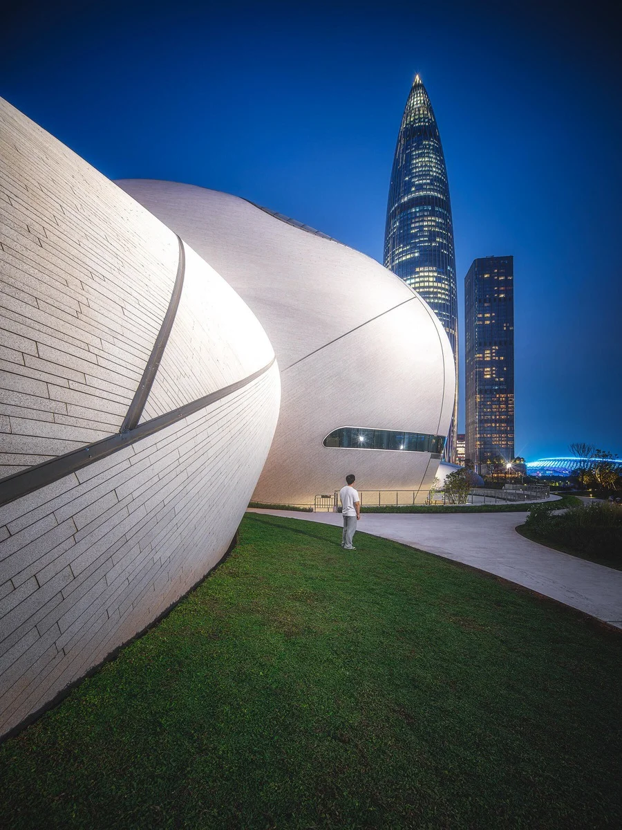 A person stands on a grassy slope facing the curvilinear stone facade of Shenzhen Bay Cultural Plaza at dusk, with the iconic