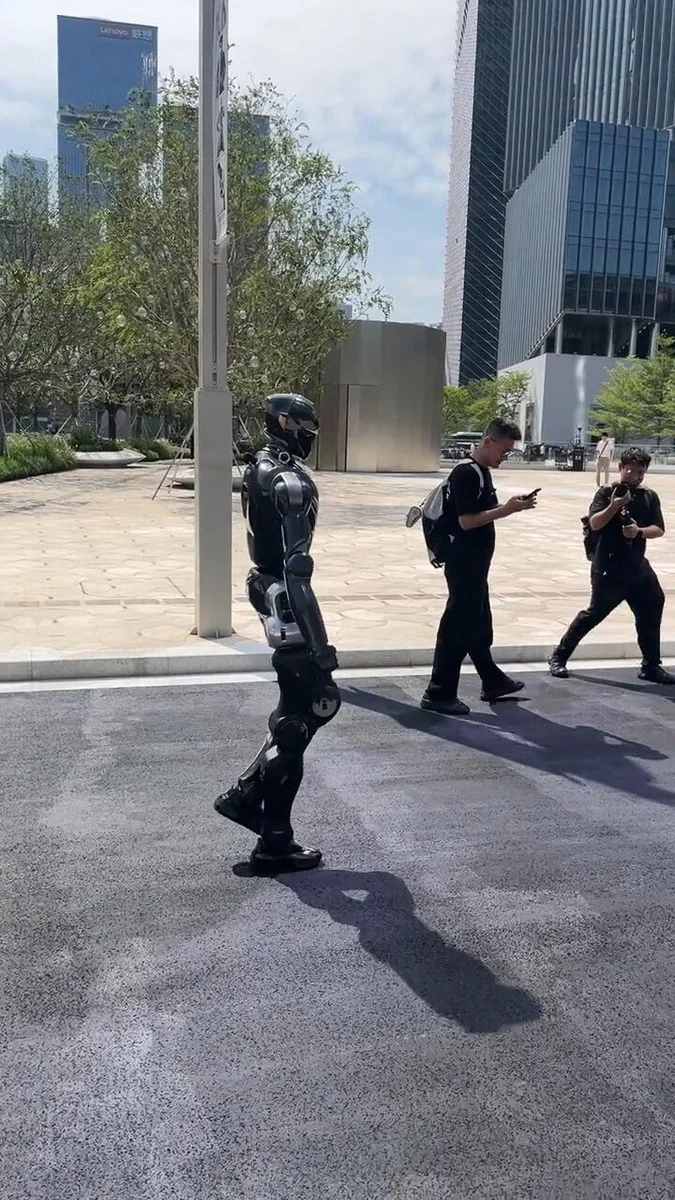 A black humanoid robot patrol unit walking on a city street in Shenzhen, with modern skyscrapers and pedestrians in the backg