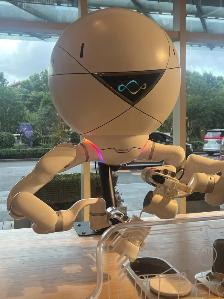 A white spherical robot with glowing arms and a triangular display is making coffee at a café counter, viewed through a windo